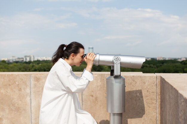 Young tourist woman standing on building rooftop looking through telescope