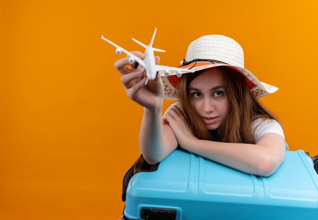 Young traveler girl wearing hat stretching model plane  and putting arm on suitcase  on isolated orange wall with copy space