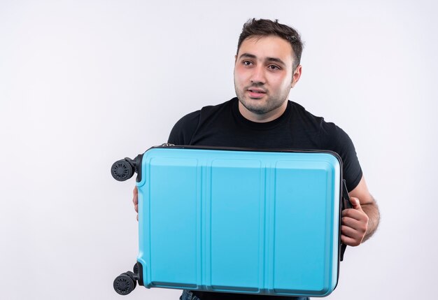 Young traveler man in black t-shirt holding suitcase looking aside puzzled standing over white wall