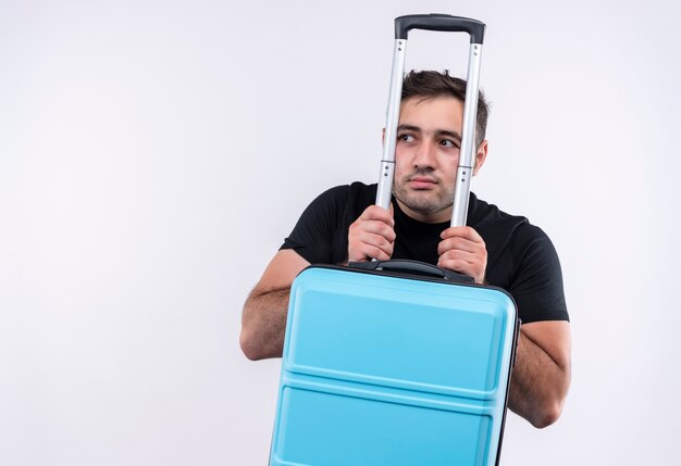 Young traveler man in black t-shirt holding suitcase looking aside worried with fear expression standing over white wall