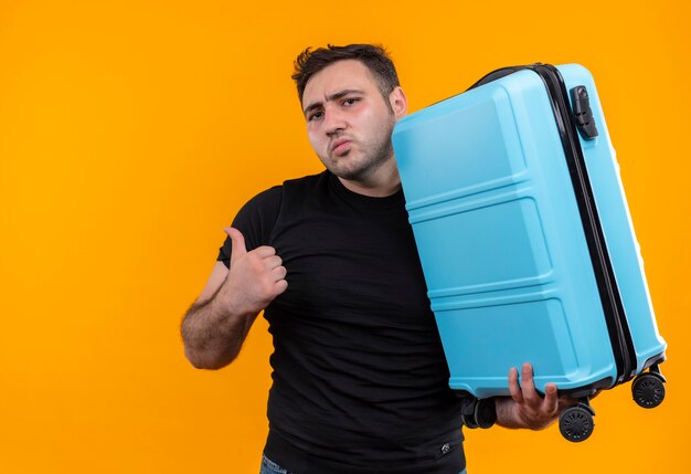 Young traveler man in black t-shirt holding suitcase looking confused pointing back with thumb standing over orange wall