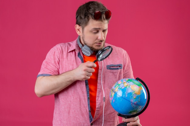 Free photo young traveler man with headphones looking at globe through magnifying glass standing over pink background