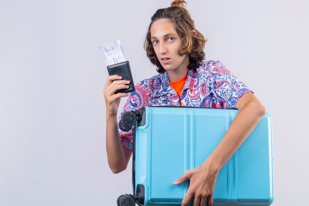 Young traveler man with suitcase holding air tickets looking confused standing over white background