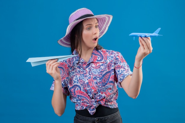 Free photo young traveler woman in summer hat holding paper and toy airplanes looking surprised and amazed over blue wall