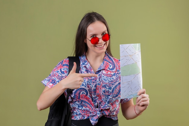 Free photo young traveler woman wearing red sunglasses and with backpack holding map pointing with index finger to it smiling cheerfully over blue wall