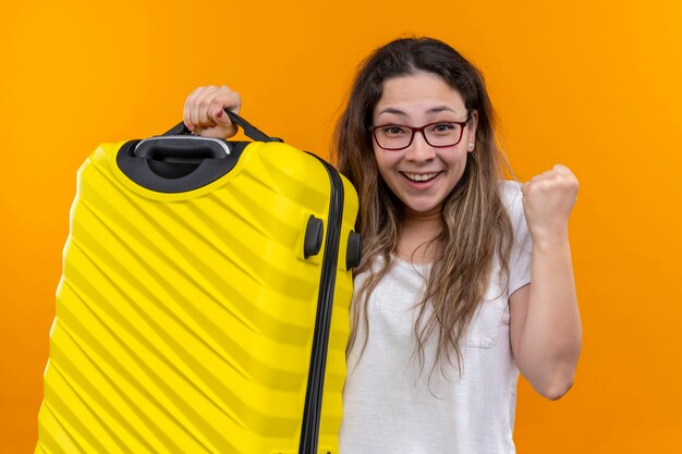 Young traveler woman in white t-shirt  holding suitcase excited and happy clenching fist standing over orange wall