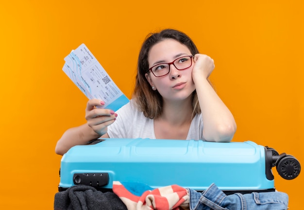 Young traveler woman in white t-shirt   holding suitcase full of clothes and air tickets leaning head on hand looking aside with pensive expression on face standing