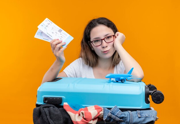 Young traveler woman in white t-shirt   holding suitcase full of clothes and air tickets leaning head on hand looking smiling standing