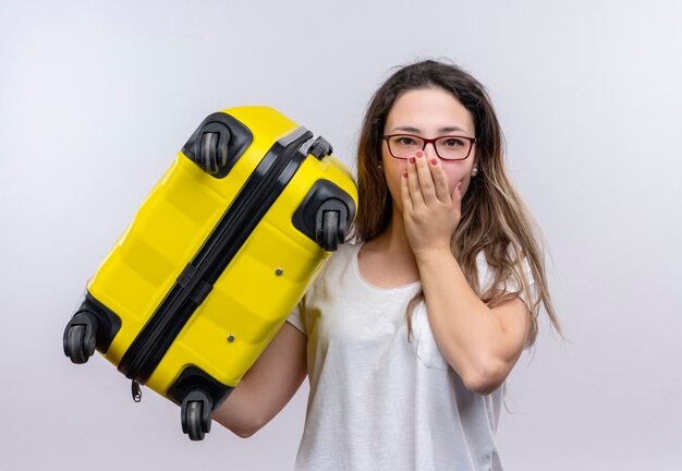 Young traveler woman in white t-shirt holding suitcase looking amazed and surprised covering mouth with hand standing over white wall