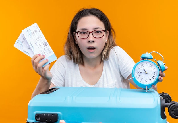 Free photo young traveler woman in white t-shirt standing with  suitcase full of clothes holding  air tickets and alarm clock looking surprised and amazed over orange wall