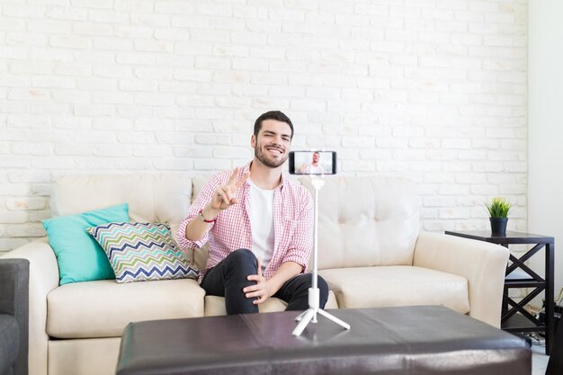 Young vlogger showing victory sign on smartphone while streaming online at home