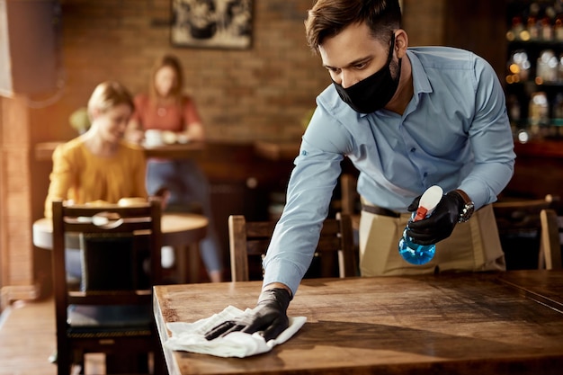 Free photo young waiter wearing protective face mask while cleaning tables while working in a cafe