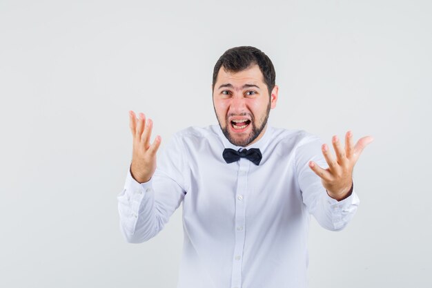 Young waiter in white shirt shouting with hands in aggressive manner and looking agitated , front view.