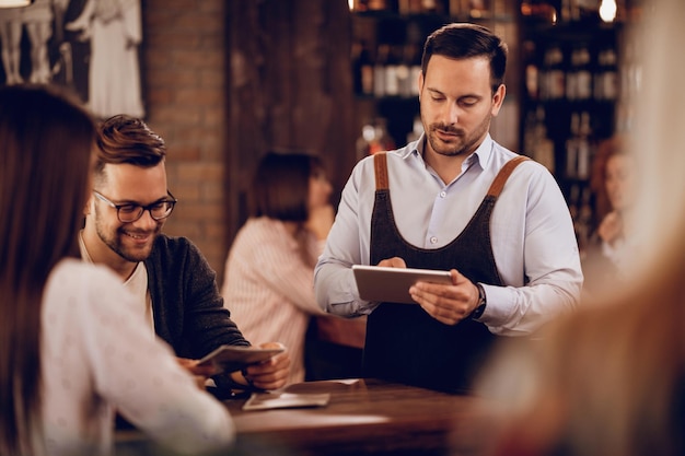 Young waiter writing order on touchpad while serving guests in a bar