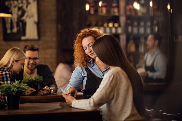 Young waitress assisting to a woman with choosing the order from a menu in a bar