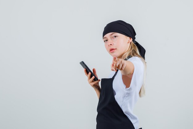 Young waitress in uniform and apron pointing while holding smartphone .