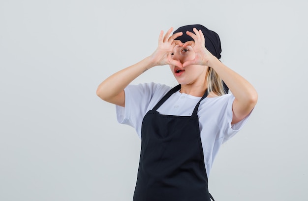 Young waitress in uniform and apron showing heart gesture on eye