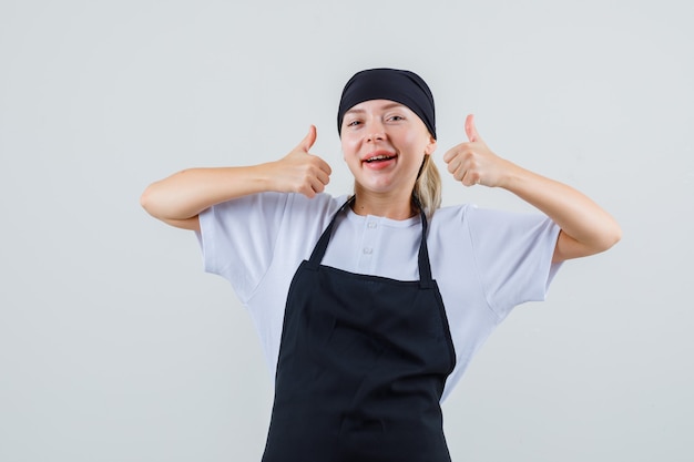 Young waitress in uniform and apron showing thumbs up and looking merry