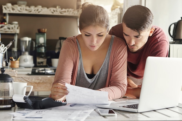 Young wife looking at piece of paper with serious look, sitting at kitchen table with laptop, calculator and documents