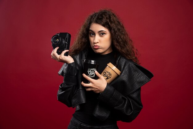 Young woman in all black outfit holding cups and a camera.