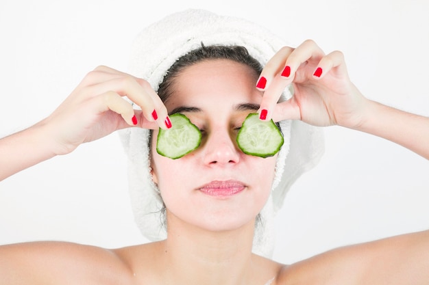 Young woman applying cucumber slices over her eyes against white background