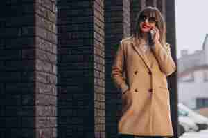 Free photo young woman in beige coat using phone in the street