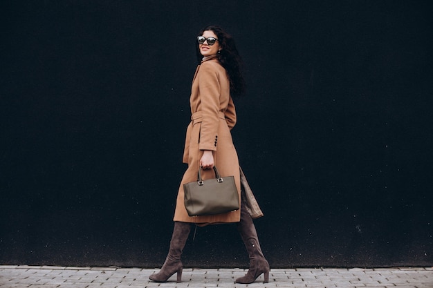 Young woman in beige coat walking outdoors on black wall background