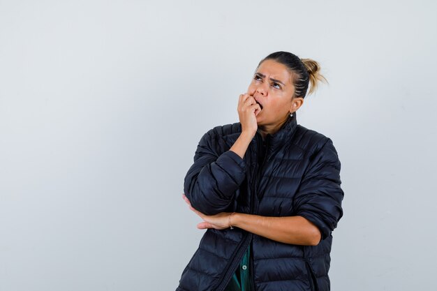 Young woman biting fist while looking up in puffer jacket and looking pensive. front view.