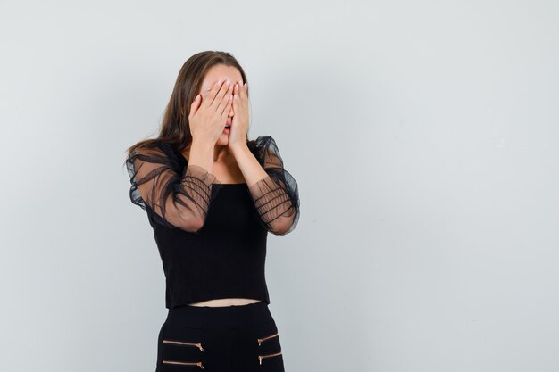 Young woman in black blouse and black pants covering face with both hands and looking stressed 