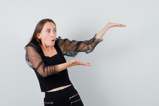 Young woman in black blouse and black pants holding hands as presenting something and looking surprised 