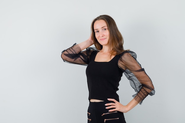 Free photo young woman in black blouse and black pants holding one hand on waist and another hand behind head and looking optimistic