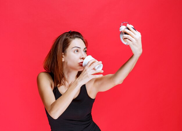 Young woman in black singlet holding an alarm clock while drinking a cup of tea