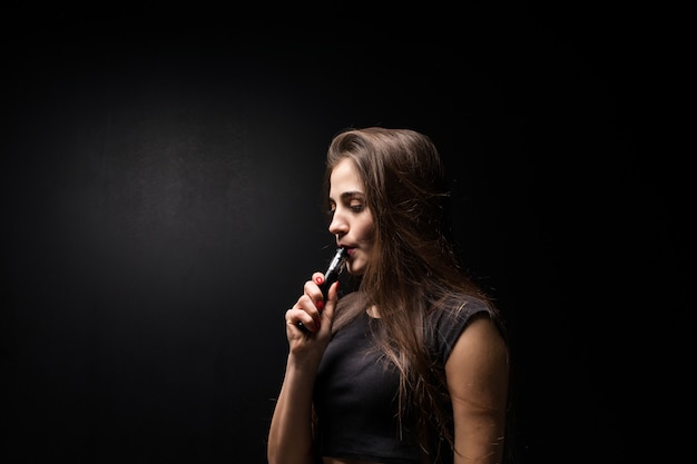 Young woman in black smokes an electronic cigarette on dark wall