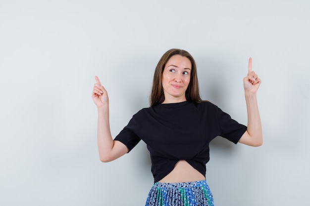 Young woman in black t-shirt and blue skirt pointing up with index fingers and looking attractive