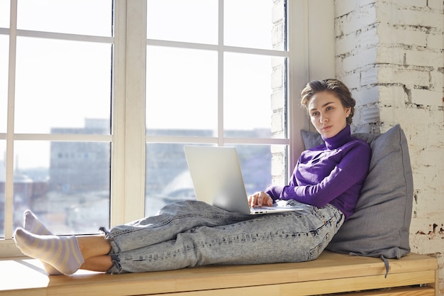 Free photo young woman blogger in stylish clothes sitting on windowsill with pillow behind her back, having pensive thoughtful look while working on new article or post for her blog on social media, using laptop