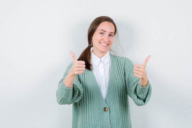 Free photo young woman in blouse, cardigan showing double thumbs up and looking merry , front view.