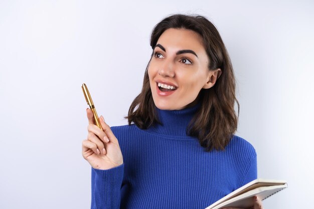 Young woman in a blue golf turtleneck on a white background pensive with a notebook in her hand thinks about ideas goals for the year