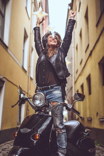 Free photo young woman in blue jeans and black jacket standing on moto scooter with hands up.