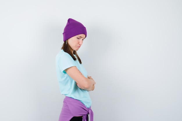 Free photo young woman in blue t-shirt, purple beanie standing arms crossed, looking down and looking dismal , front view.