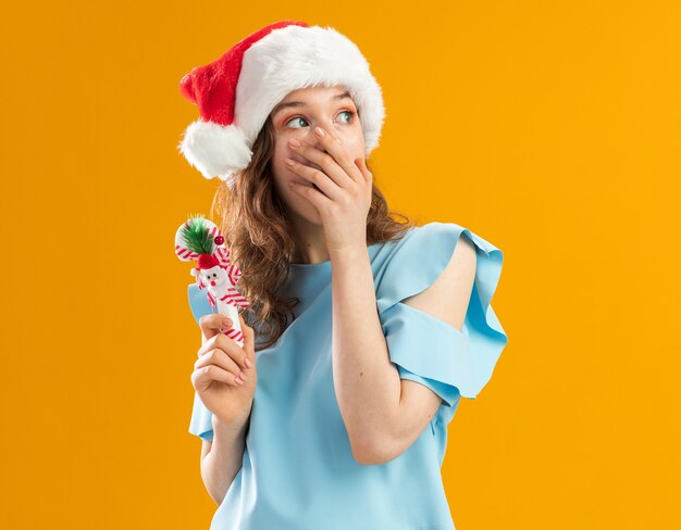 Young woman in blue top and santa hat holding christmas candy cane looking aside being shocked covering mouth with hand 