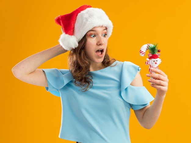 Free photo young woman in blue top and santa hat holding christmas candy cane looking at it amazed with hand on her head