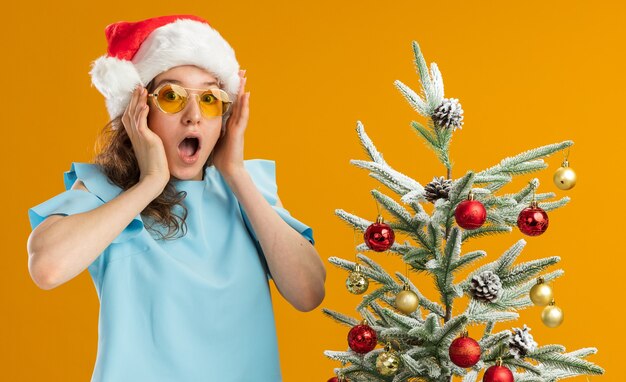 Young woman in blue top and santa hat wearing yellow glasses   amazed and surprised standing next to a christmas tree over orange wall