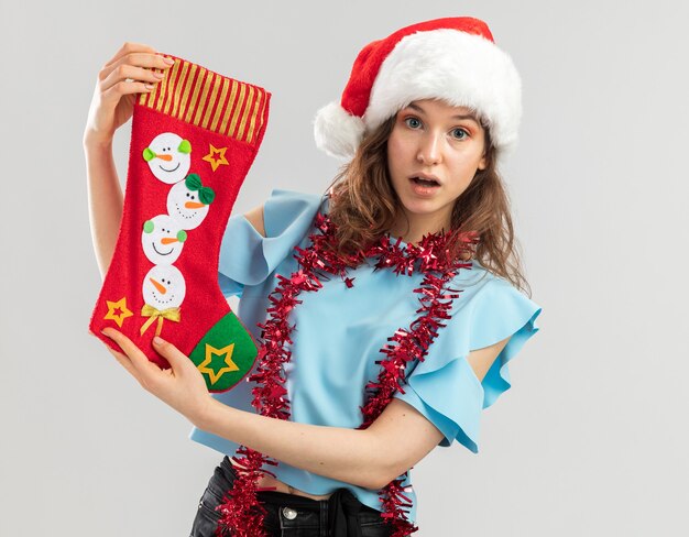 Young woman in blue top and santa hat with tinsel around her neck holding christmas stocking  surprised standing over white  wall