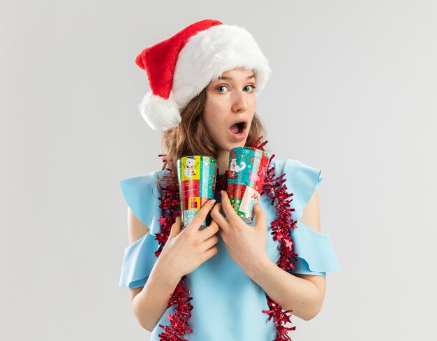 Young woman in blue top and santa hat with tinsel around her neck holding colorful paper cups looking surprised 