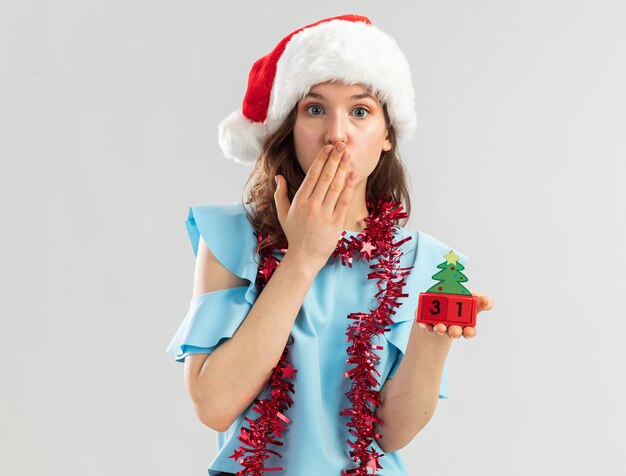 Young woman in blue top and santa hat with tinsel around her neck holding toy cubes with happy ney year date looking being shocked covering mouth with hand 