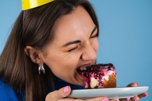 Free photo young woman on a blue wall celebrates a birthday, holds a piece of cake, happy, bites the cake