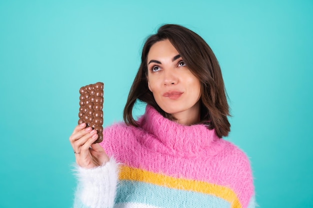 Free photo a young woman in a bright multi-colored sweater on blue enjoys every piece of a delicious bar of airy milk chocolate