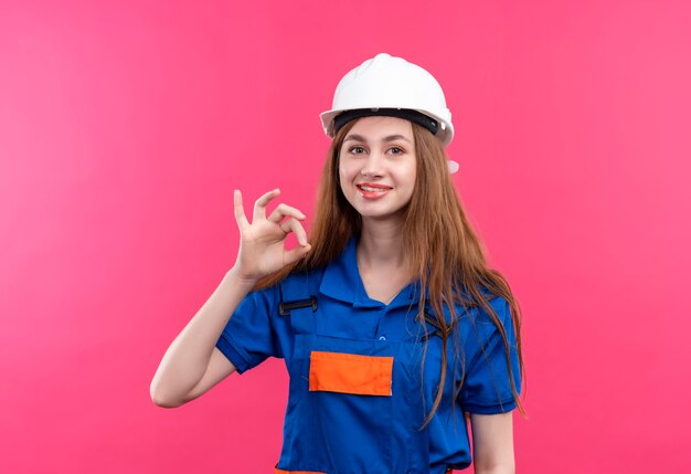 Young woman builder worker in construction uniform and safety helmet happy and positive smiling showing ok sign standing over pink wall