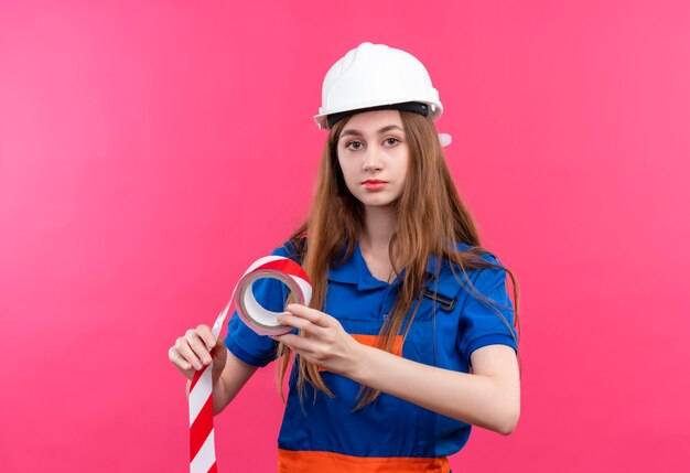 Young woman builder worker in construction uniform and safety helmet holding scotch tape looking with serious face standing over pink wall