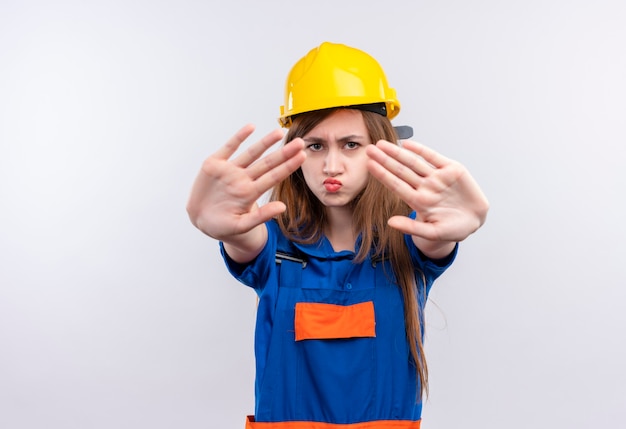 Young woman builder worker in construction uniform and safety helmet looking displeased standing with open hands making stop sign  over white wall